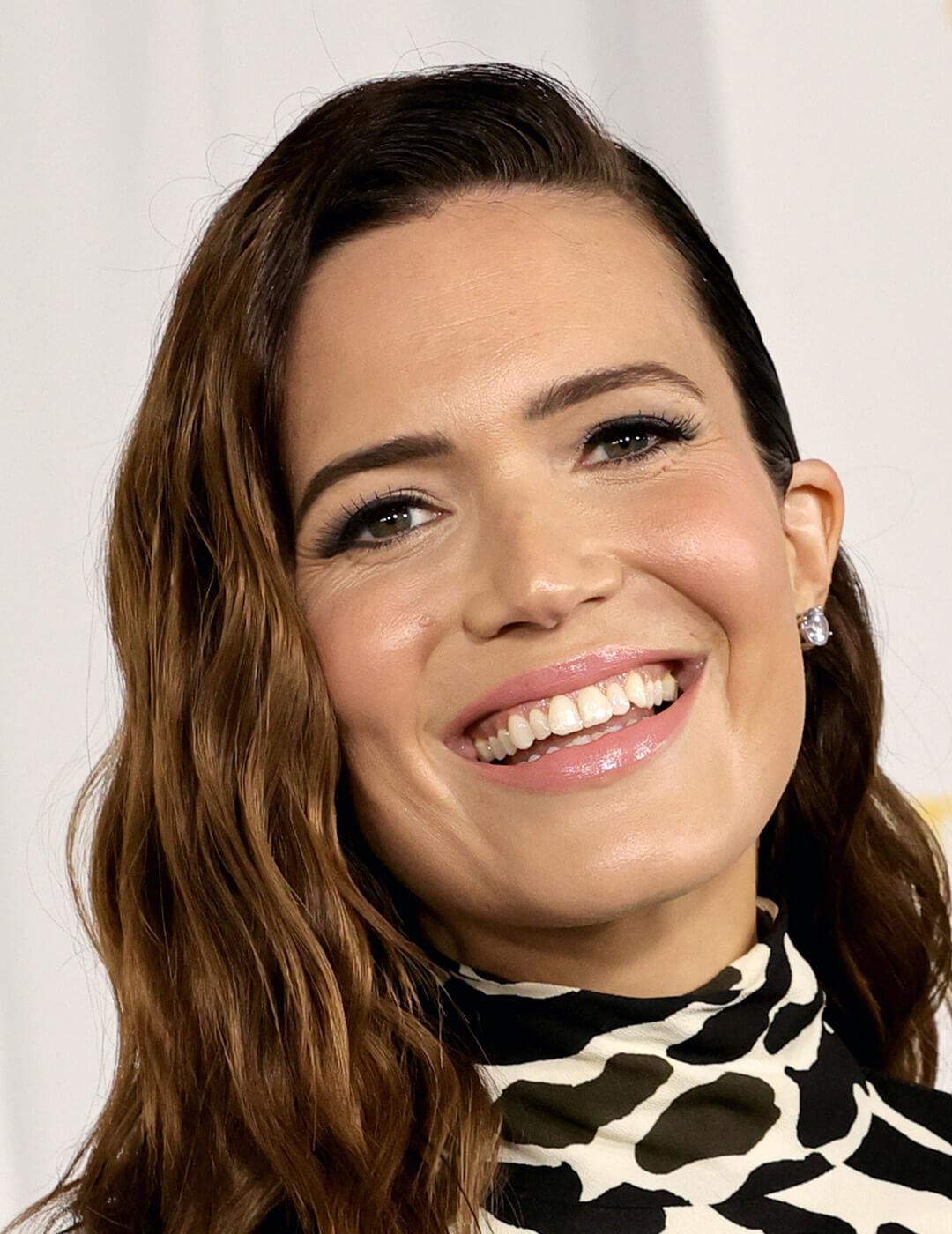 Close-up of Mandy Moore smiling at the camera rocking light pink gloss wearing a printed black and white dress complemented by her diamond earrings