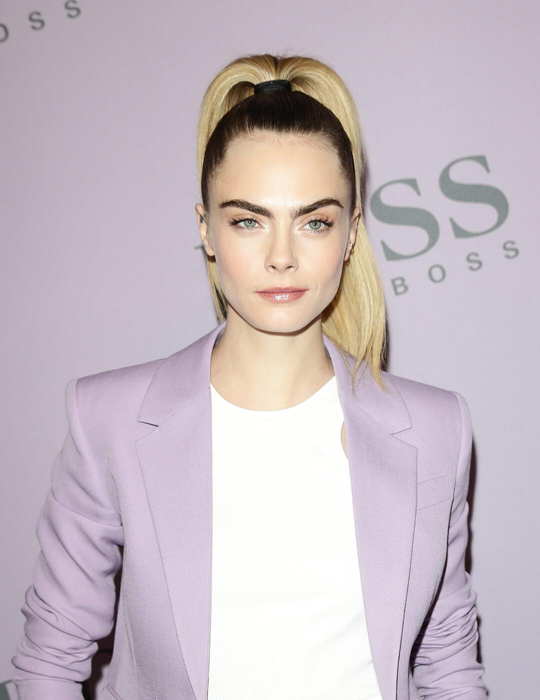 A photo of Cara Delevingne wearing a purple suit paired in white top with her blond hair styled in a simple classic chic ponytail