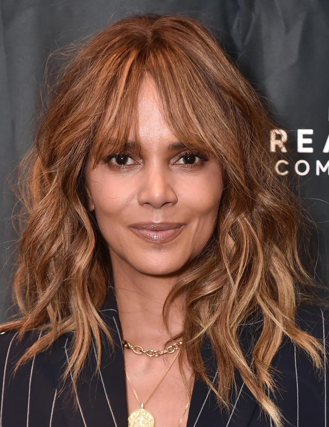 A photo of Halle Berry with dark blonde highlight extentions wearing a black coat with white stripes and gold chain necklace