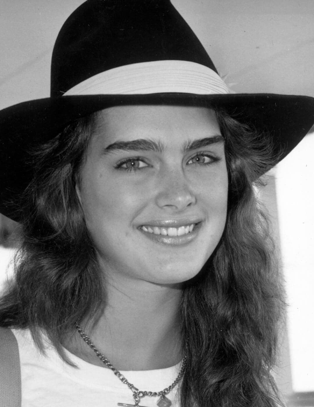A photo of Brooke Shields wearing a dark hat with a white brim, and holding a pair of sunglasses, circa 1985