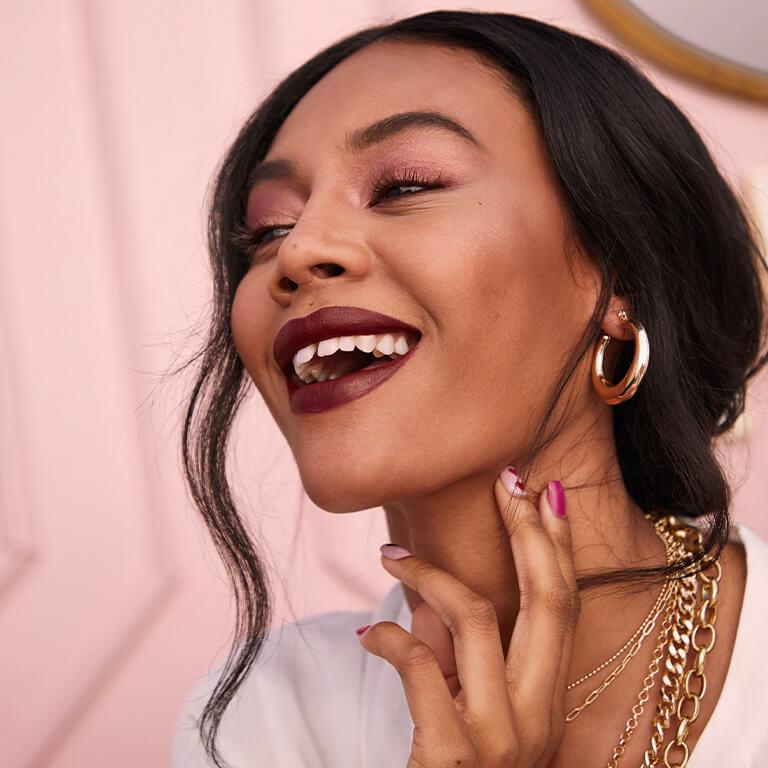 An image of a model wearing gold jewelry, rose gold eyeshadow, and dark berry lipstick smiling big and holding her neck with two fingers