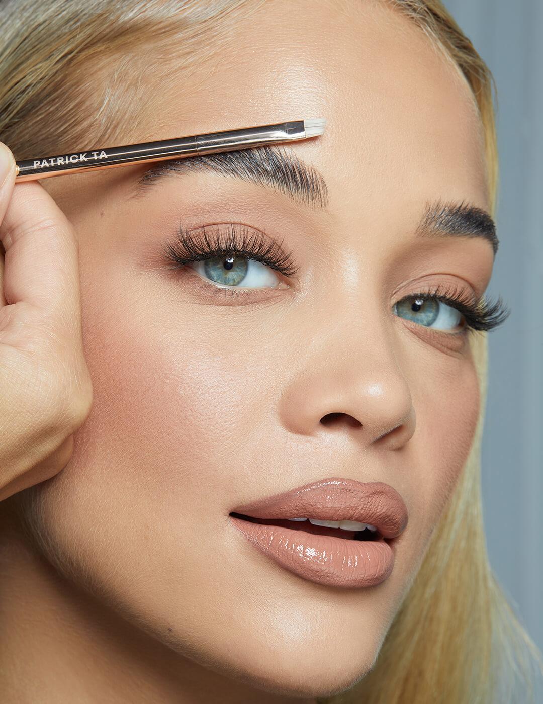 Close-up image of Jasmine Sanders with a neutral makeup look and a makeup brush touching her eyebrow