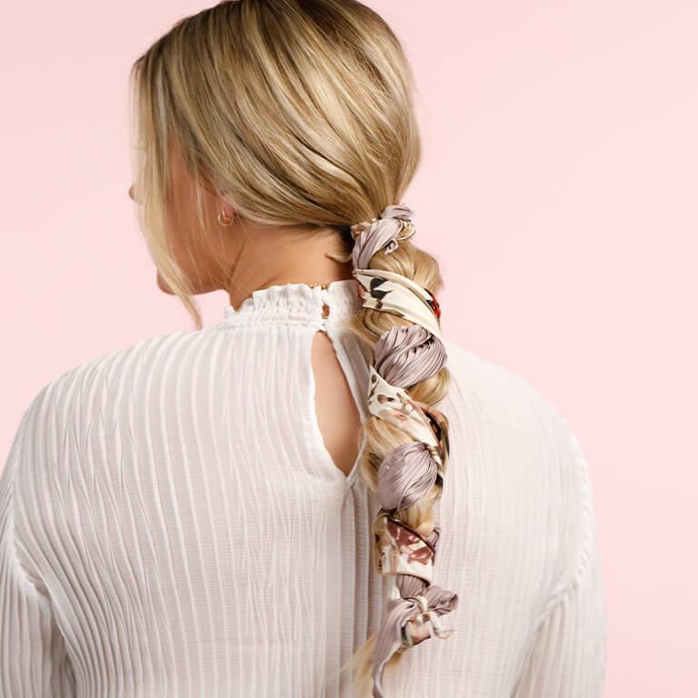 An image of a blonde model showing her braids interlaced with a white and lilac floral scarf