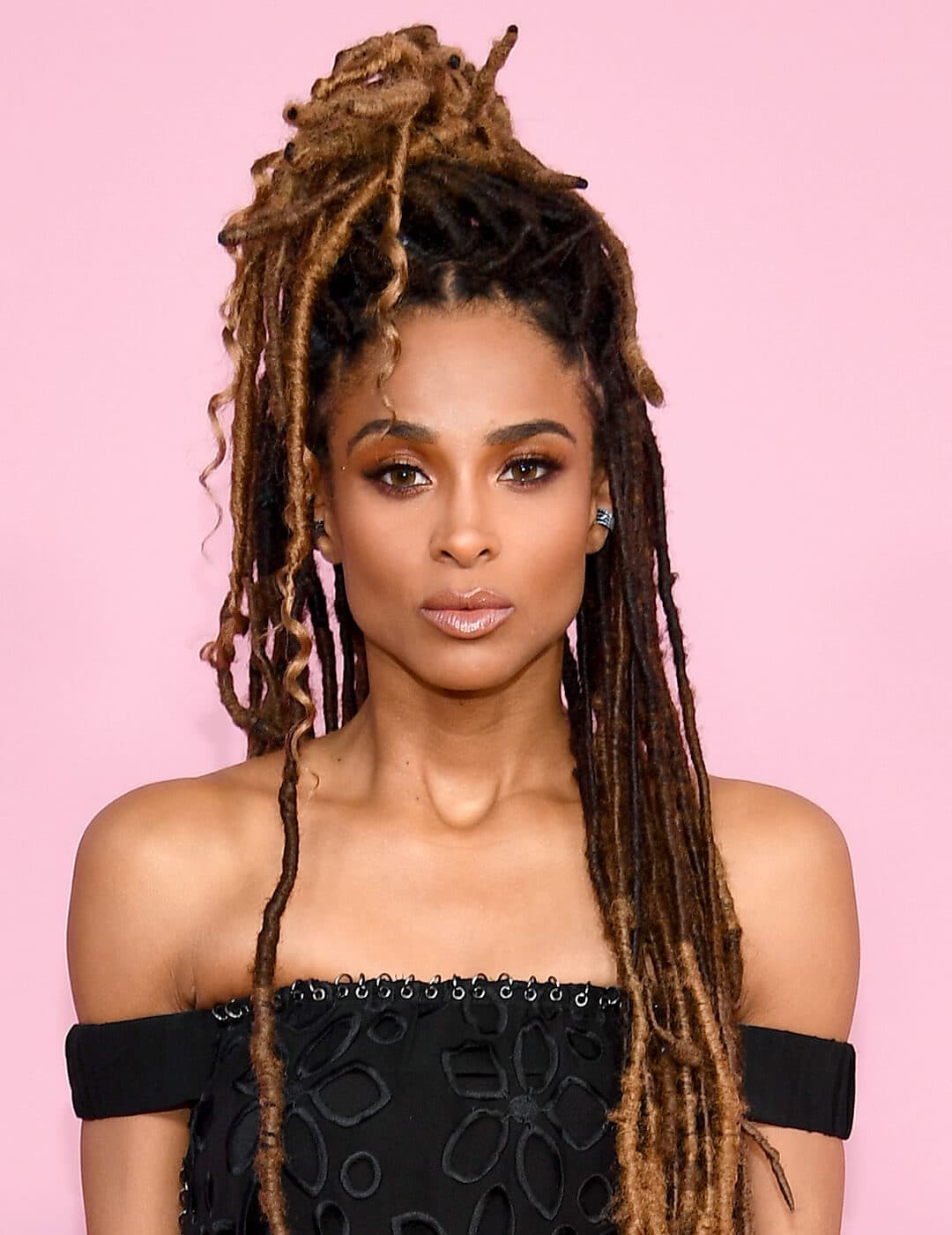 Close-up of Ciara rocking a faux locs half updo hairstyle, smoky eye makeup paired with a nude lipstick, and black dress against pink background