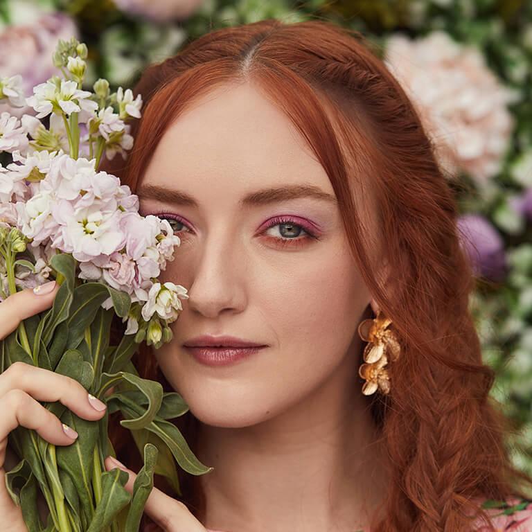 A close-up image of a red-haired model rocking a matte fuschia and peach eyeshadow look and floral earrings while covering half of her face with a bouquet of flowers