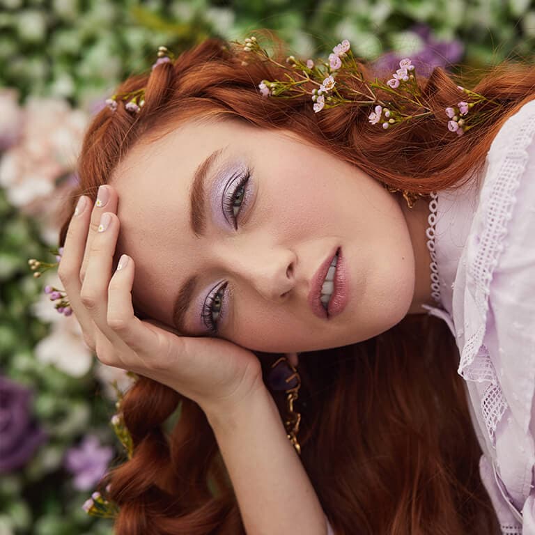 A close-up profile image of a braided red-haired model with flowers wearing a shimmery lilac purple eyeshadow look