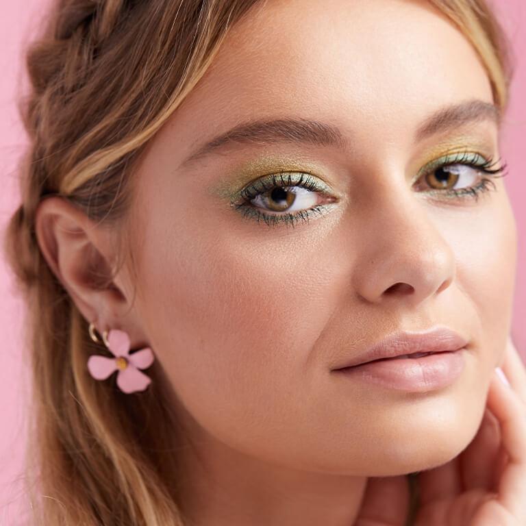 A close-up image of a model wearing a shimmery lime green and mint green eyeshadow look paired with a nude pink lipstick