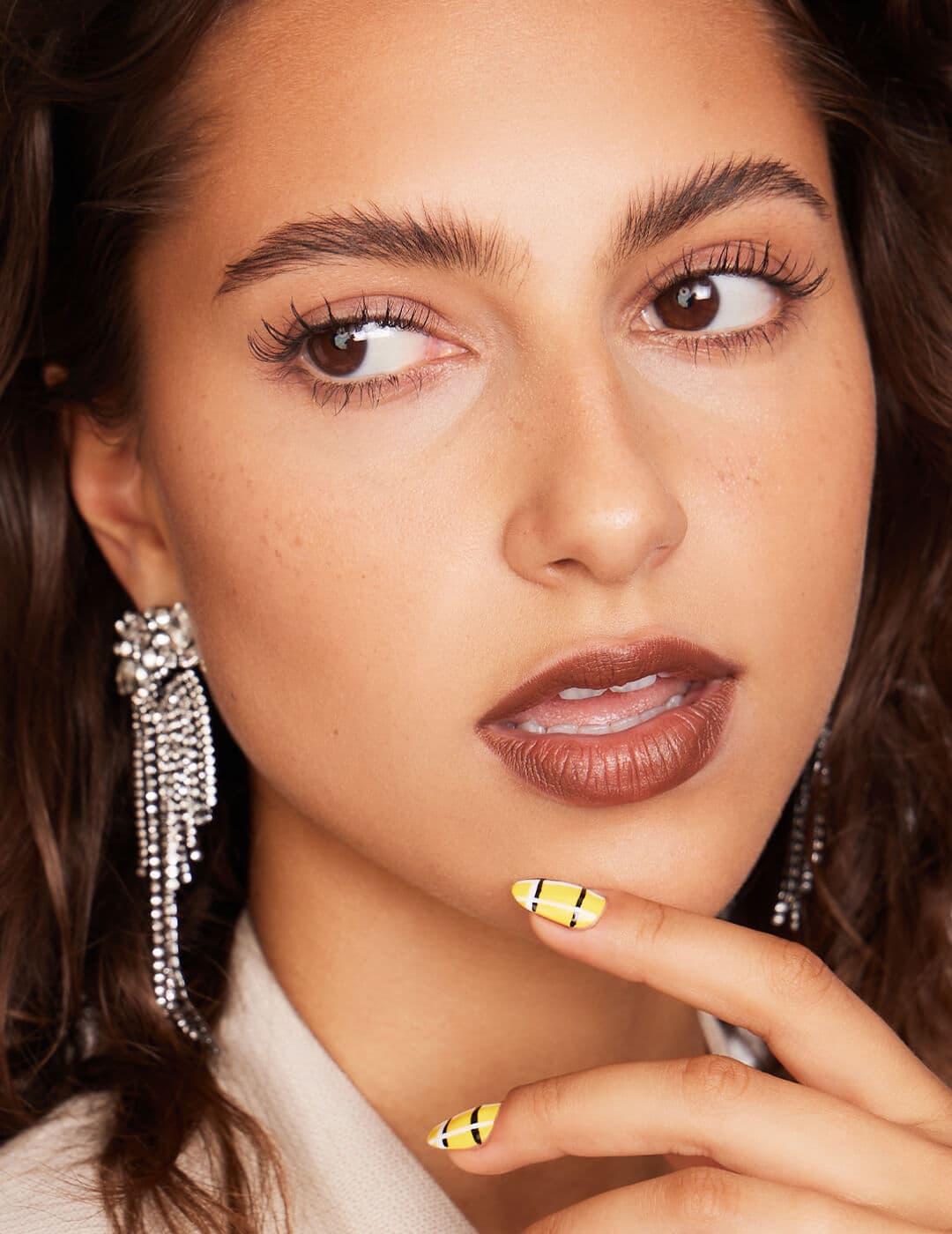 Close-up of a model rocking silver dangling earrings, yellow and black stripes nail art, and a brick lip makeup look