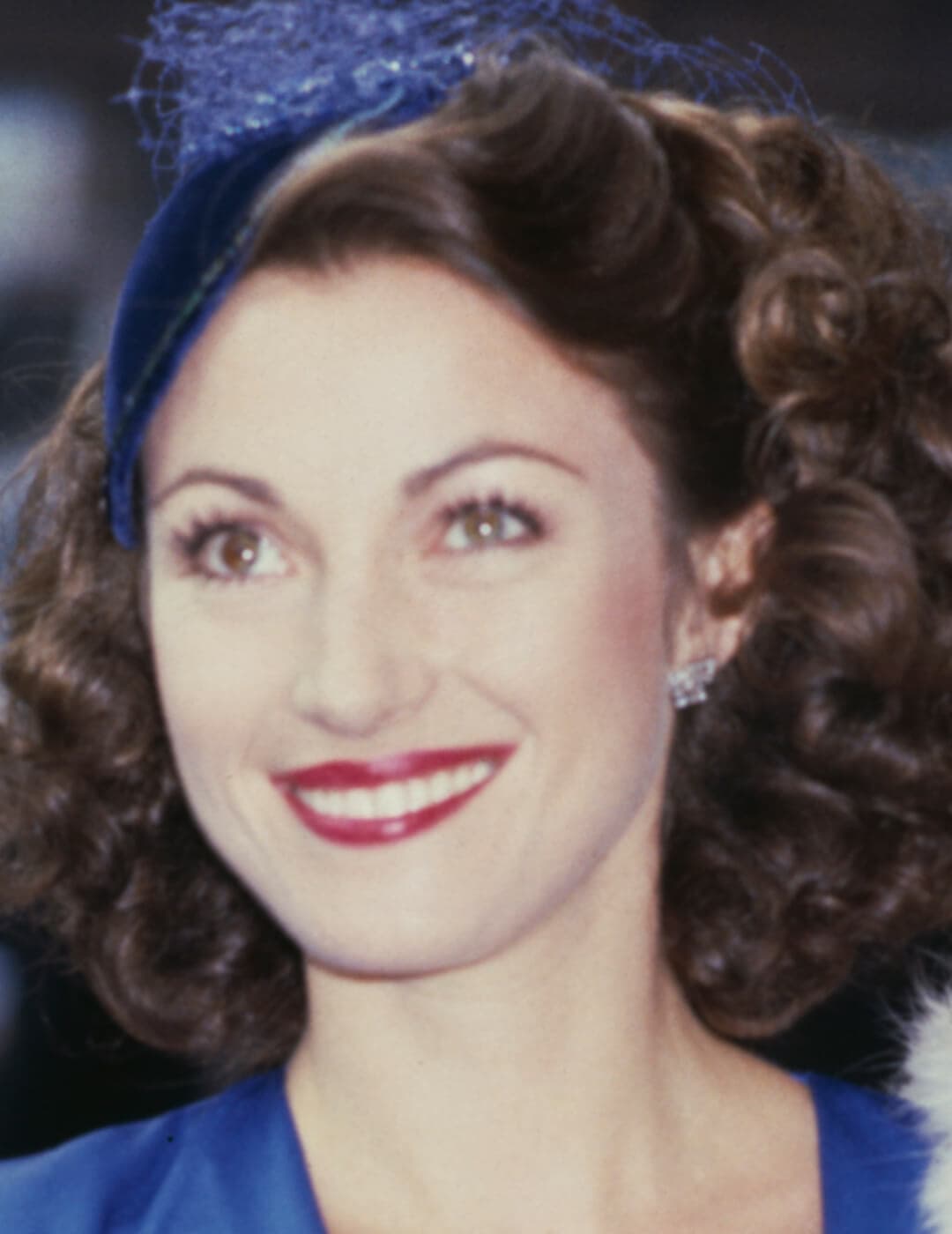 A photo of Jane Seymour wearing a blue outfit with plunging neckline and a blue fascinator, and a metallic fuchsia lip