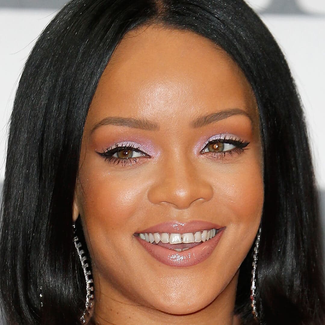 Close-up image of a smiling Rihanna wearing a shimmery lavender eyeshadow makeup look and silver dangling earrings