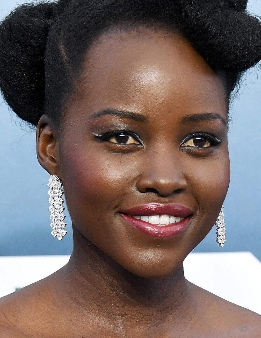 Lupita Nyong'o rocking silver dangling earrings, black and white eye makeup look, and glossy nude pink lips