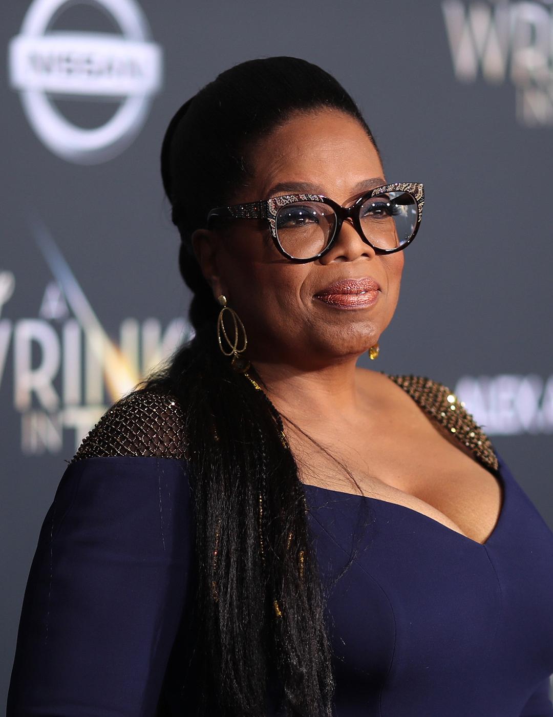 A photo of Oprah Winfrey wearing a royal blue long-sleeved dress, black cats eye glasses with her slicked-back low ponytail 