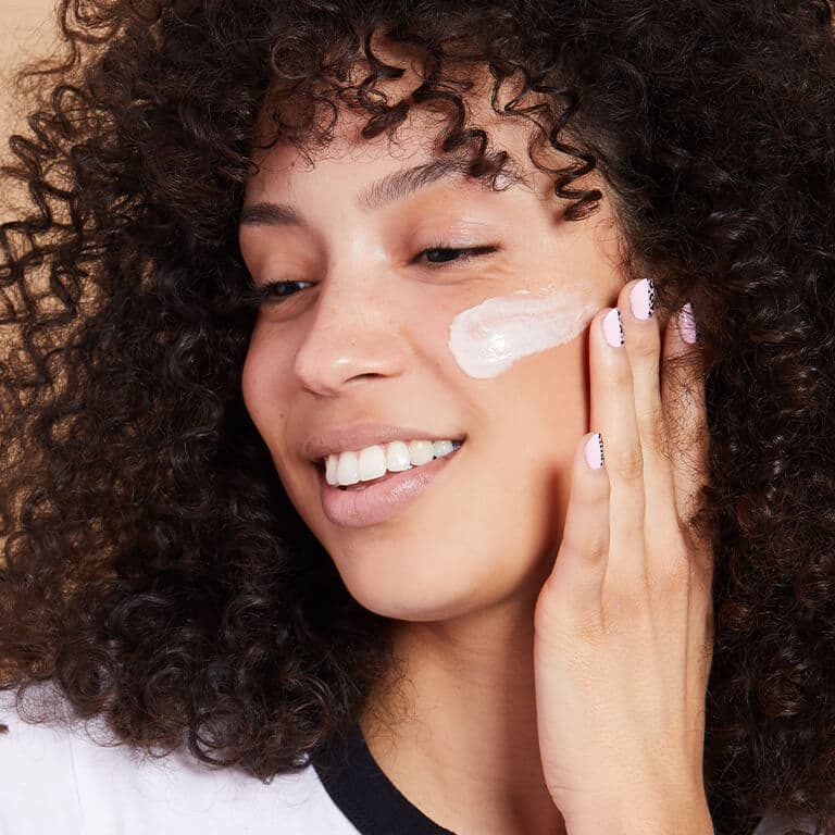 An image of a smiling model with curly hair applying cream on her cheek