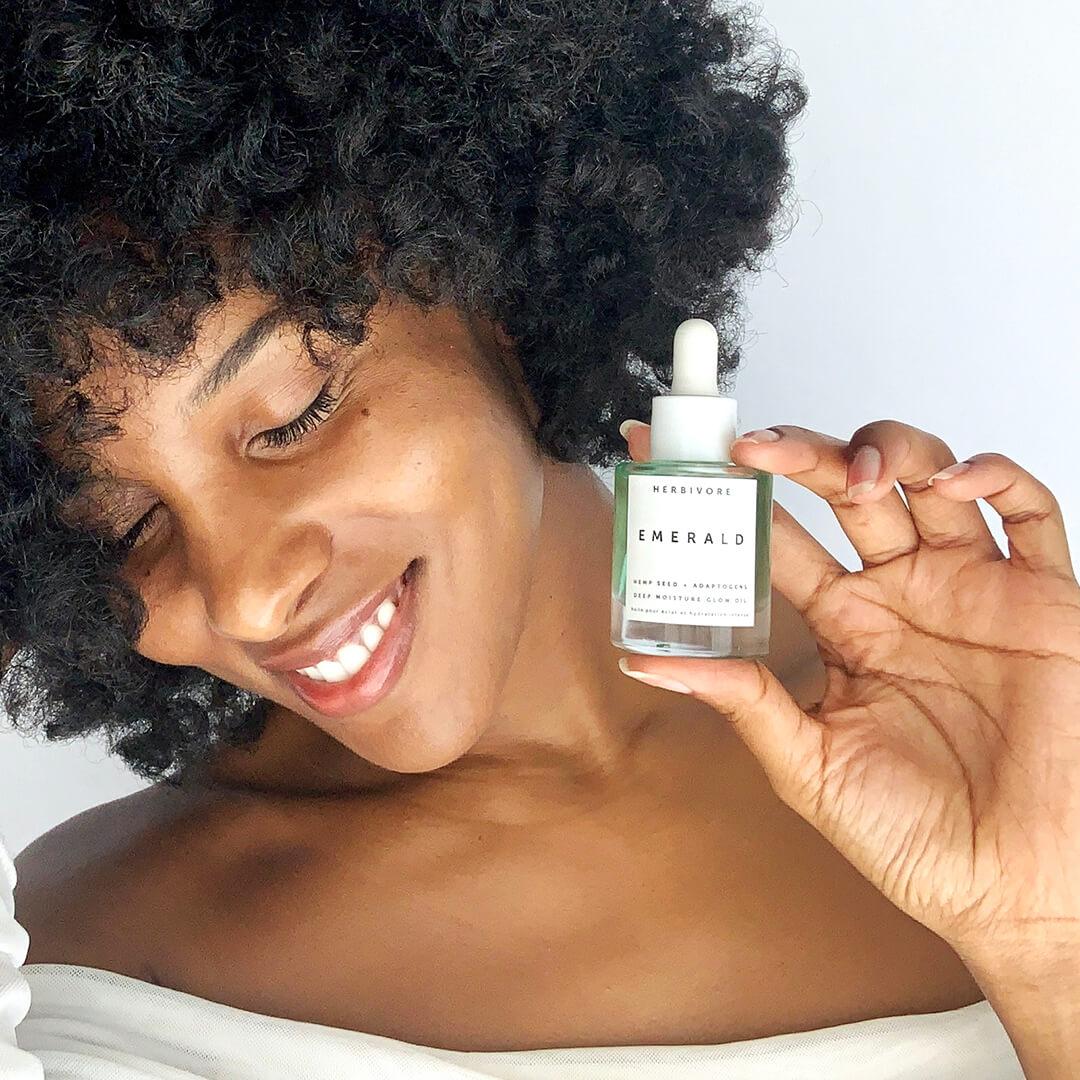 Close-up of a smiling black woman holding up the HERBIVORE Emerald Deep Moisture Glow Oil while looking down