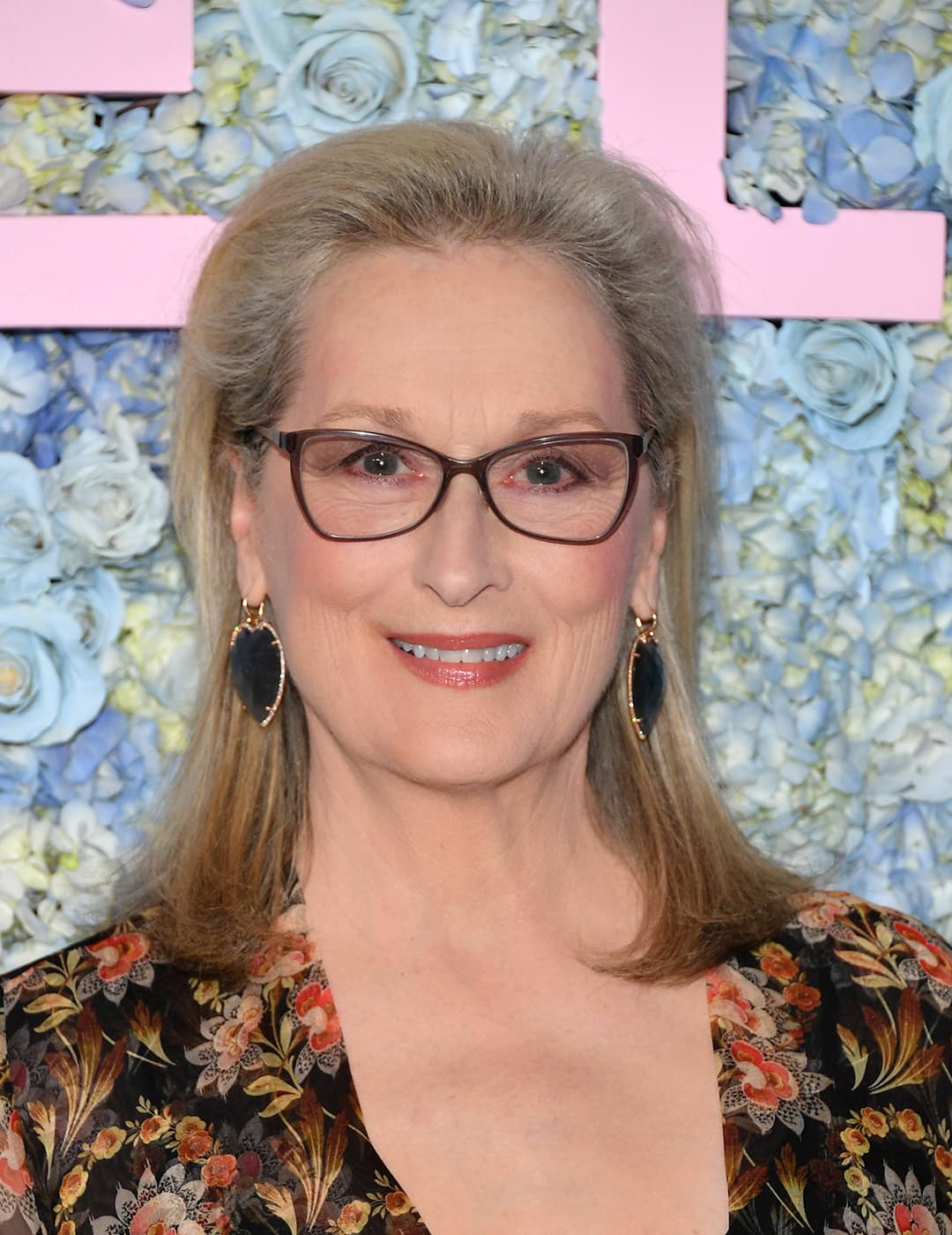 A photo of Meryl Streep wearing a classic black eyeglasses, black earing and a black floral see through dress along with her classic mid-length style hairstyle 
