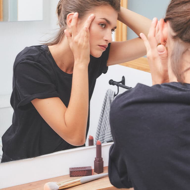 An image of a model in black shirt holding the skin on the side of her eye while looking at herself in the mirror