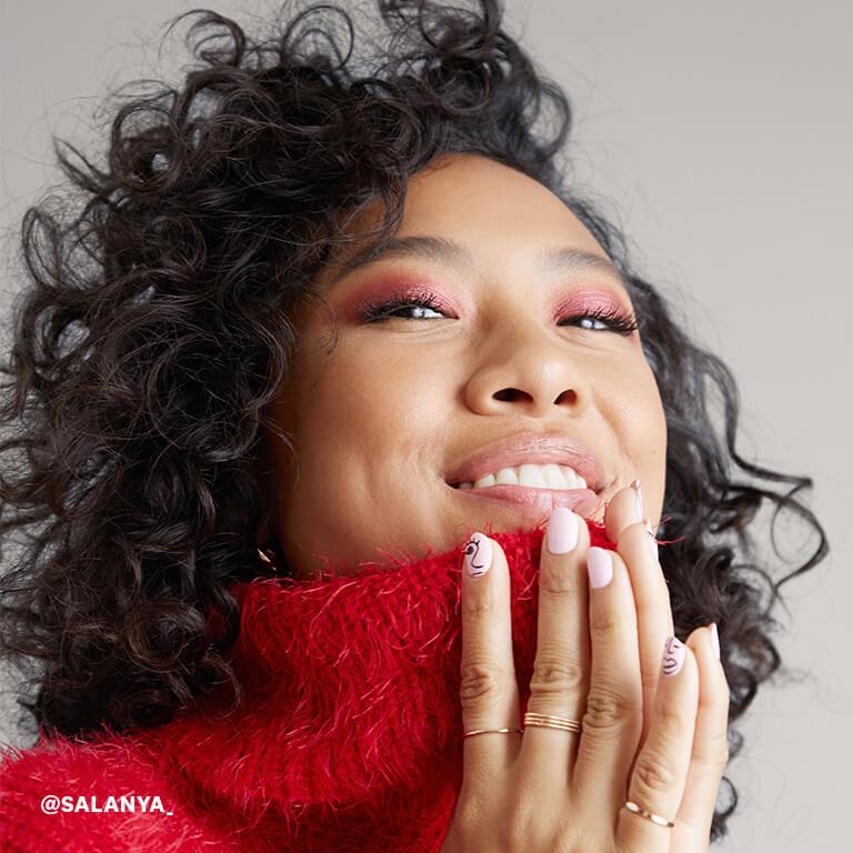 An image of a model with curly hair and hands together wearing red, shimmery eye shadow and a red, wool turtleneck sweater