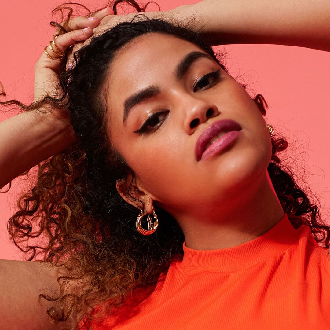 Close-up image of a model with thick, curly hair rocking a natural makeup look wearing an orange top on a red background