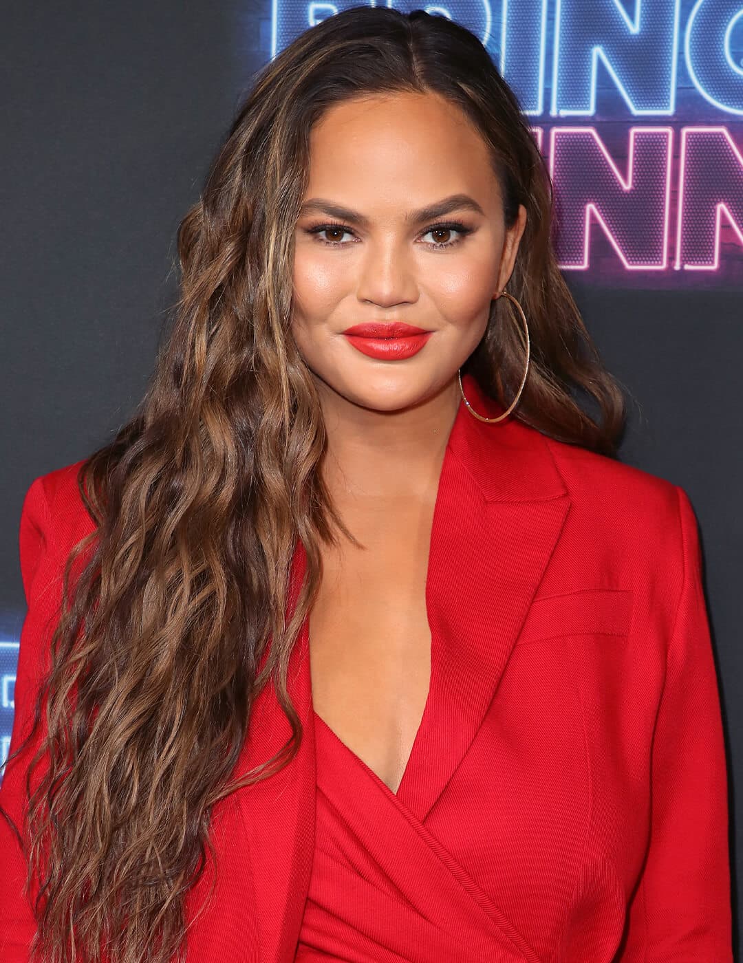 Chrissy Teigen rocking a long and wavy hairstyle with bold red lips and big hoop earrings in a red suit