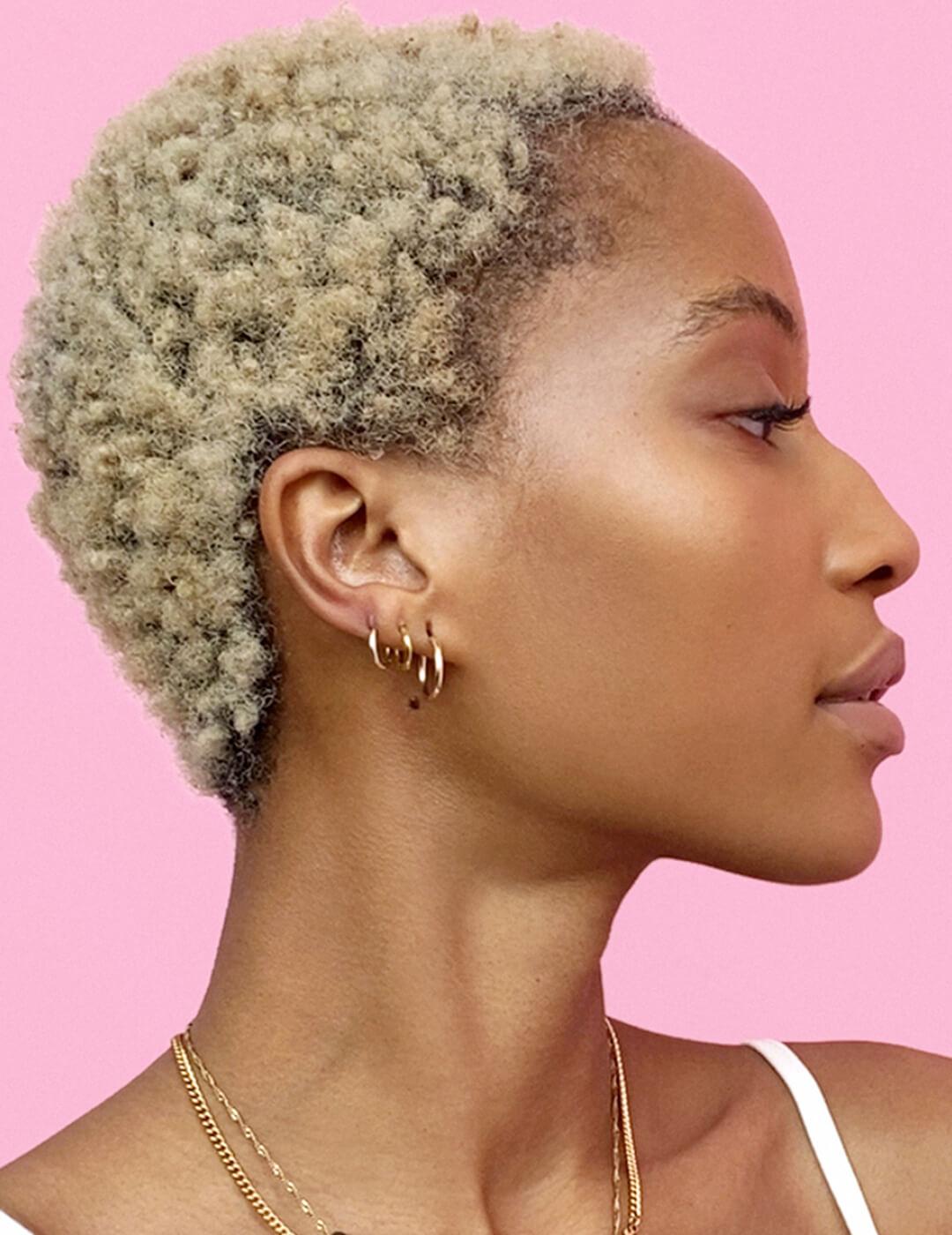 a side view photo of a woman with blonde curly hair with three earings on a pink background