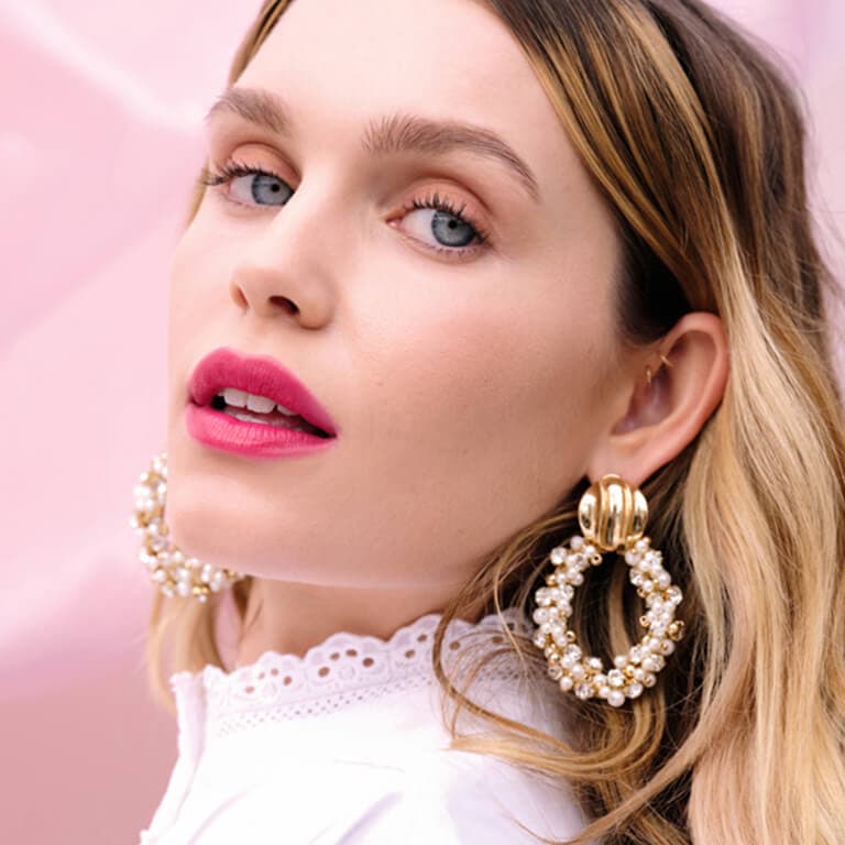 A closeup image of a model wearing nude eyeshadow and bold pink lipstick and gold and pearl hoop earrings