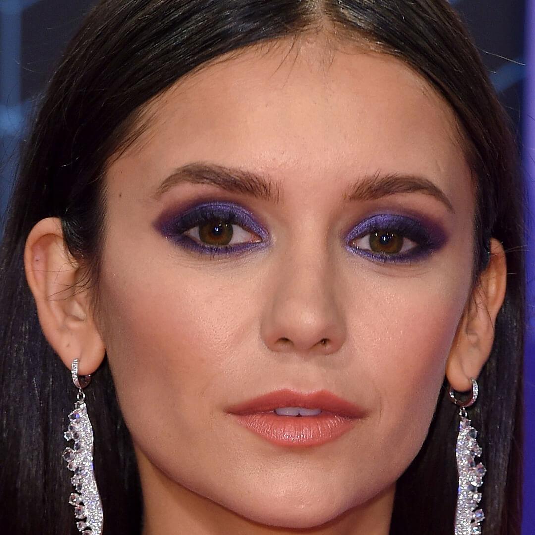 Close-up image of Nina Dobrev rocking a dark purple eye makeup look and silver feather earrings