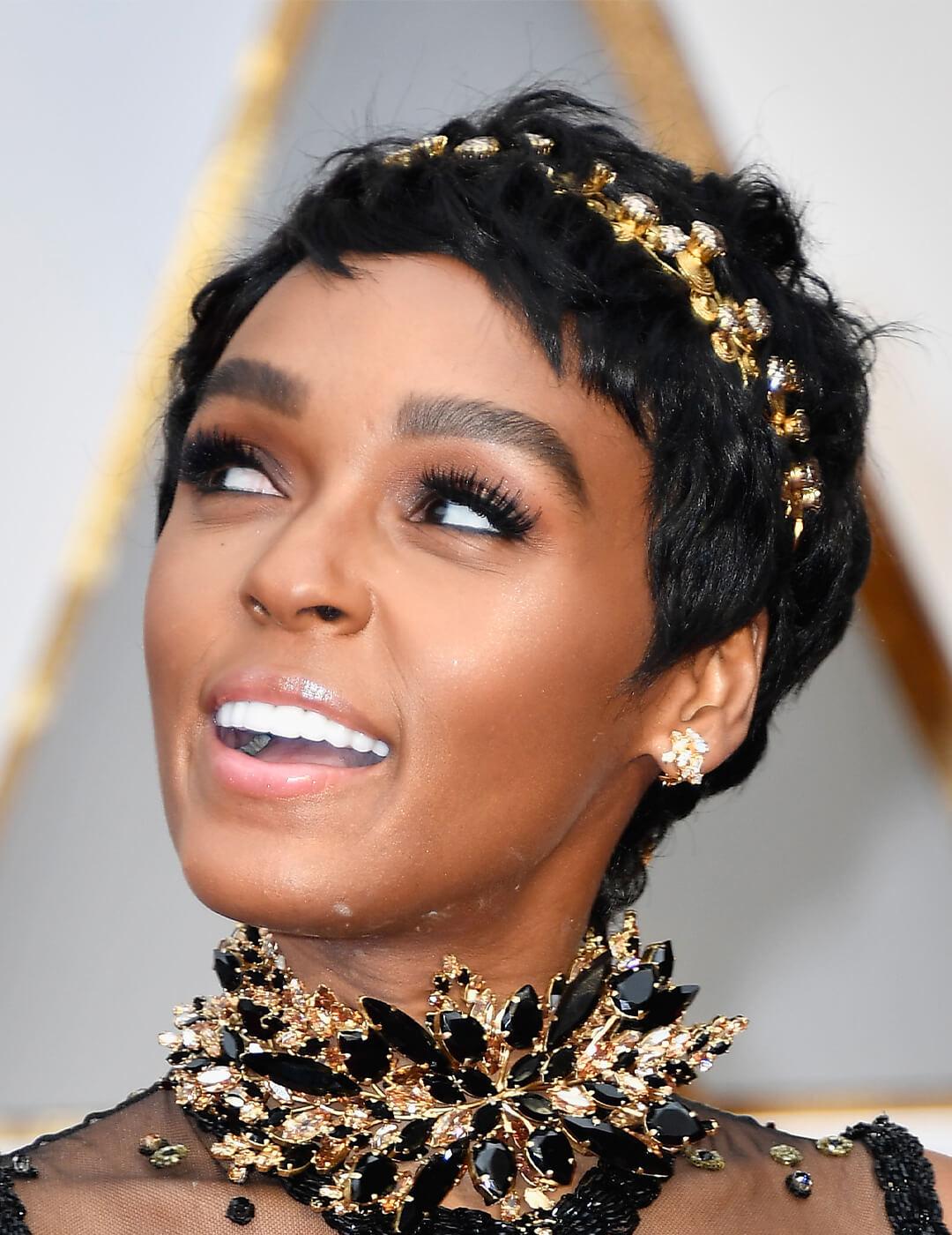 Close-up of Janelle Monae smiling while rocking a neutral makeup look, pixie cut hairstyle accented with gold and silver headband, and black and gold choker necklace