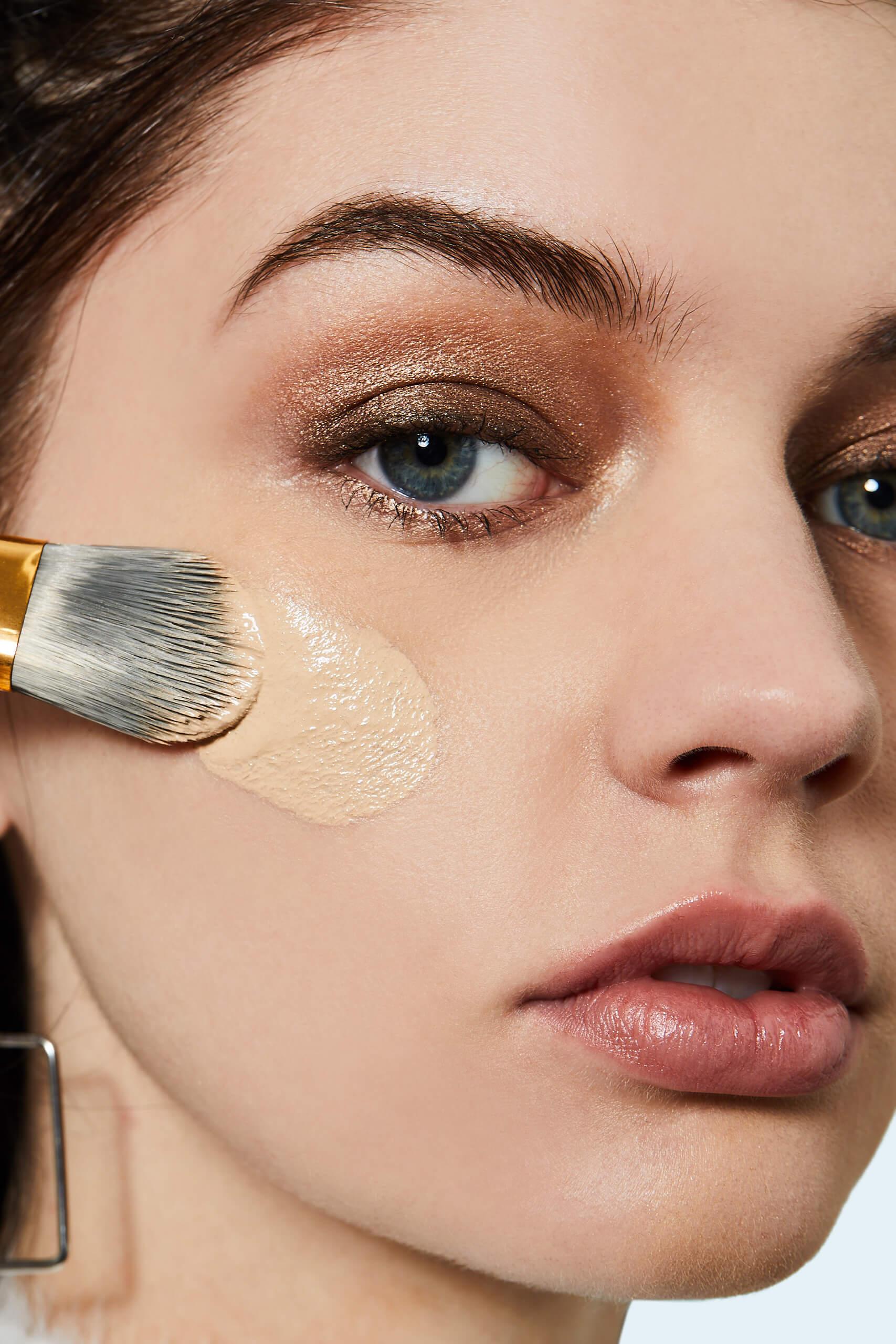Close-up image of a woman applying liquid foundation on her face with a makeup brush