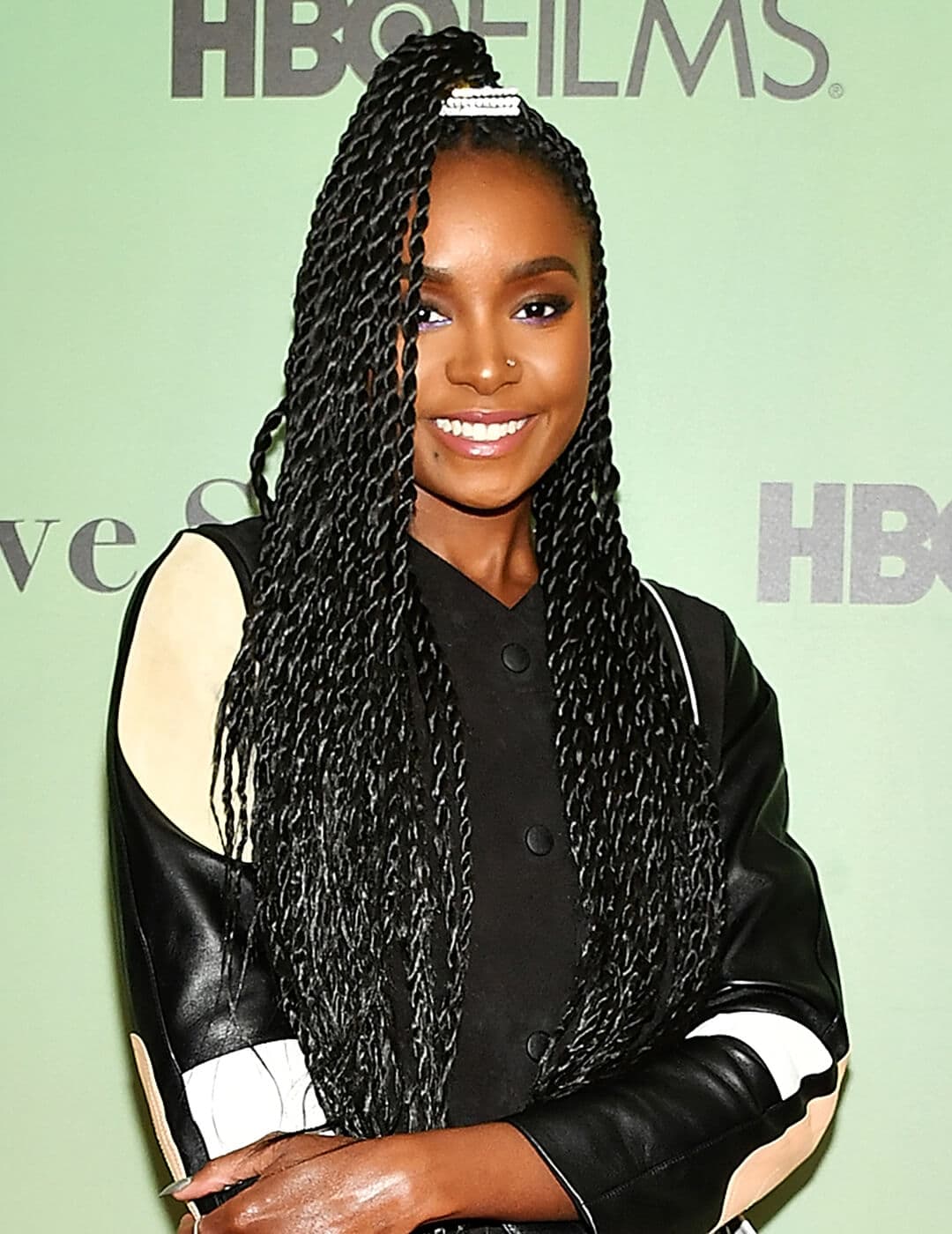 Kiki Layne rocking a high ponytail twists hairstyle, cream, white, and black leather jacket, and smoky eyeshadow makeup look paired with nude lips