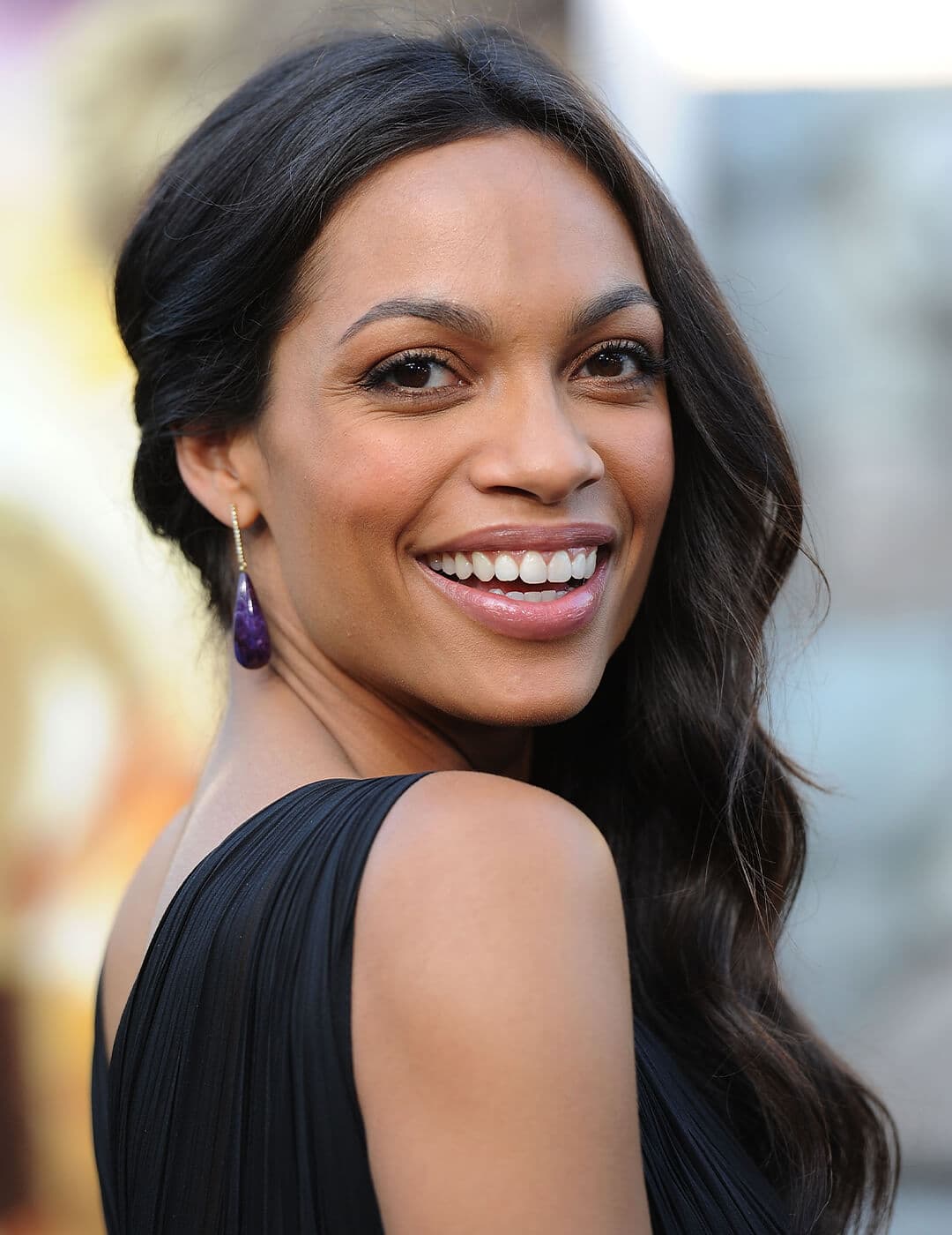 Rosario Dawson with a side swept, wavy hairstyle and in a black dress smiling