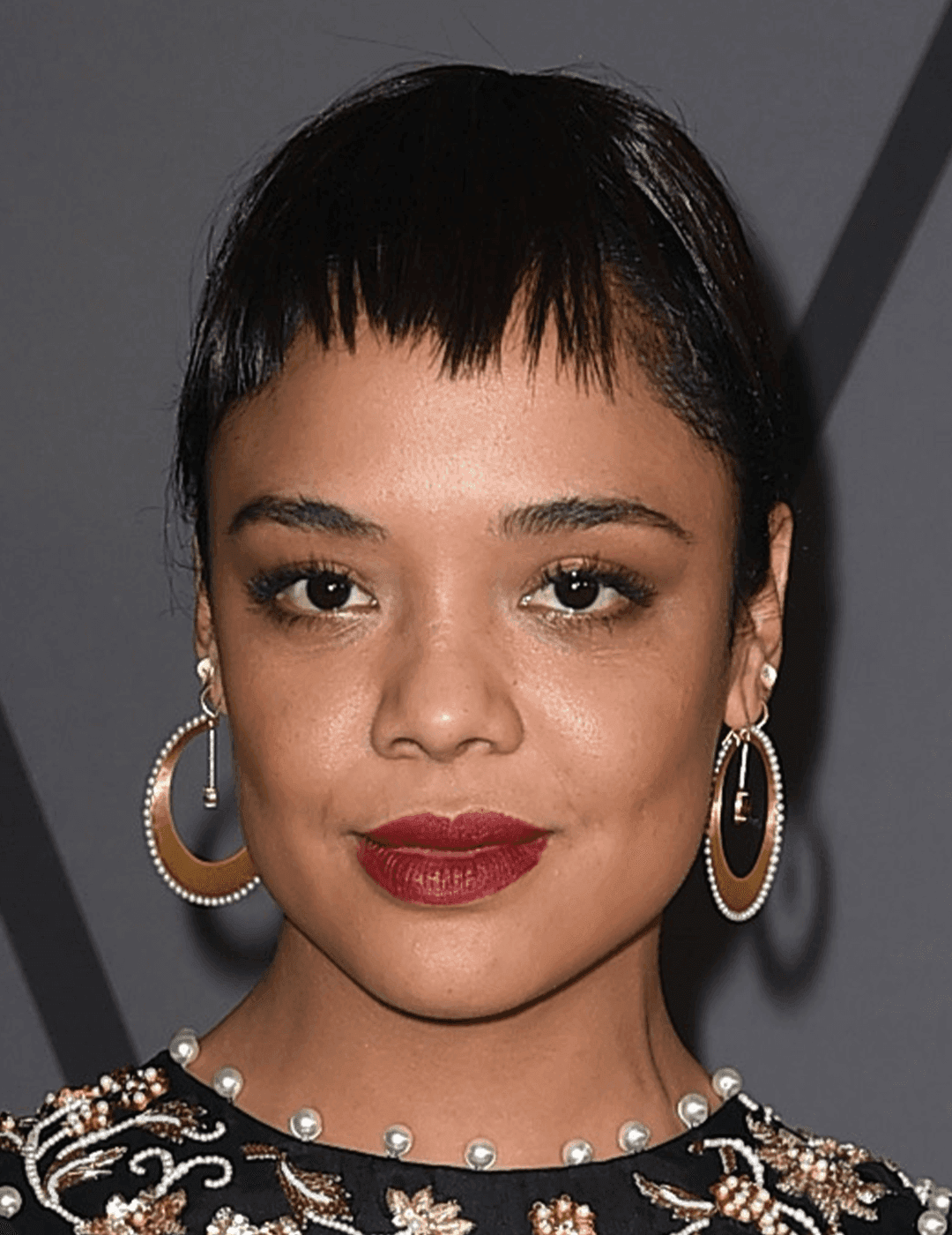 Tessa Thompson rocking a black and gold dress, neutral eye makeup with berry lips, gold hoop earrings, and a pixie cut hairstyle with micro bangs