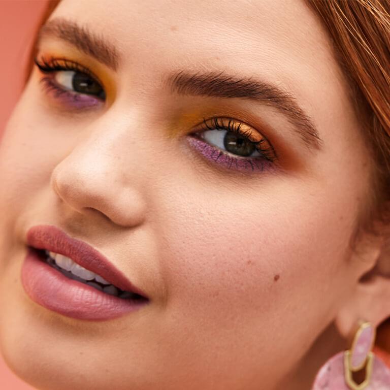 A closeup image of a model wearing bold orange and purple eyeshadow paired with a matte rose lipstick 