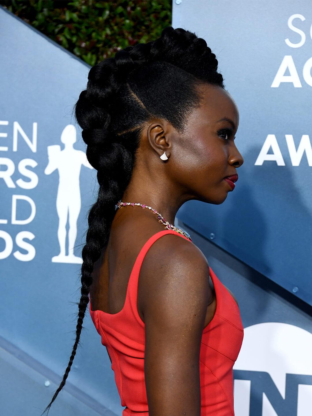 Side profile of Danai Gurira rocking a braided faux mohawk hairstyle and coral dress