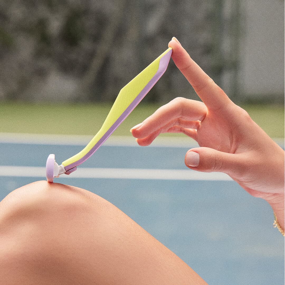 Close-up of a model holding the REFRESHMENTS Dazzling 5-Blade Razor against her knee, in a tennis court