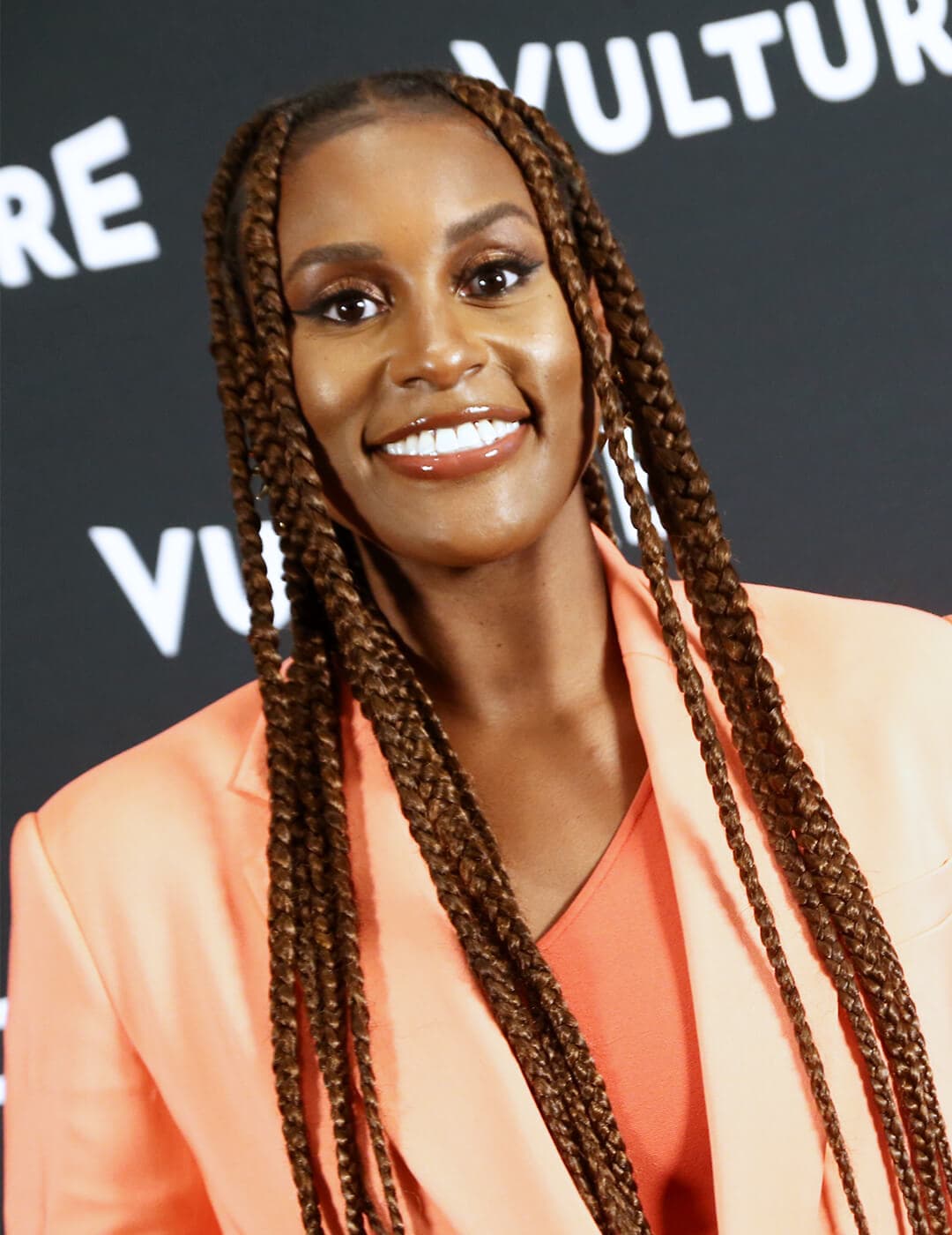 Smiling Issa Rae rocking long, braided hairstyle and orange suit on the red carpet