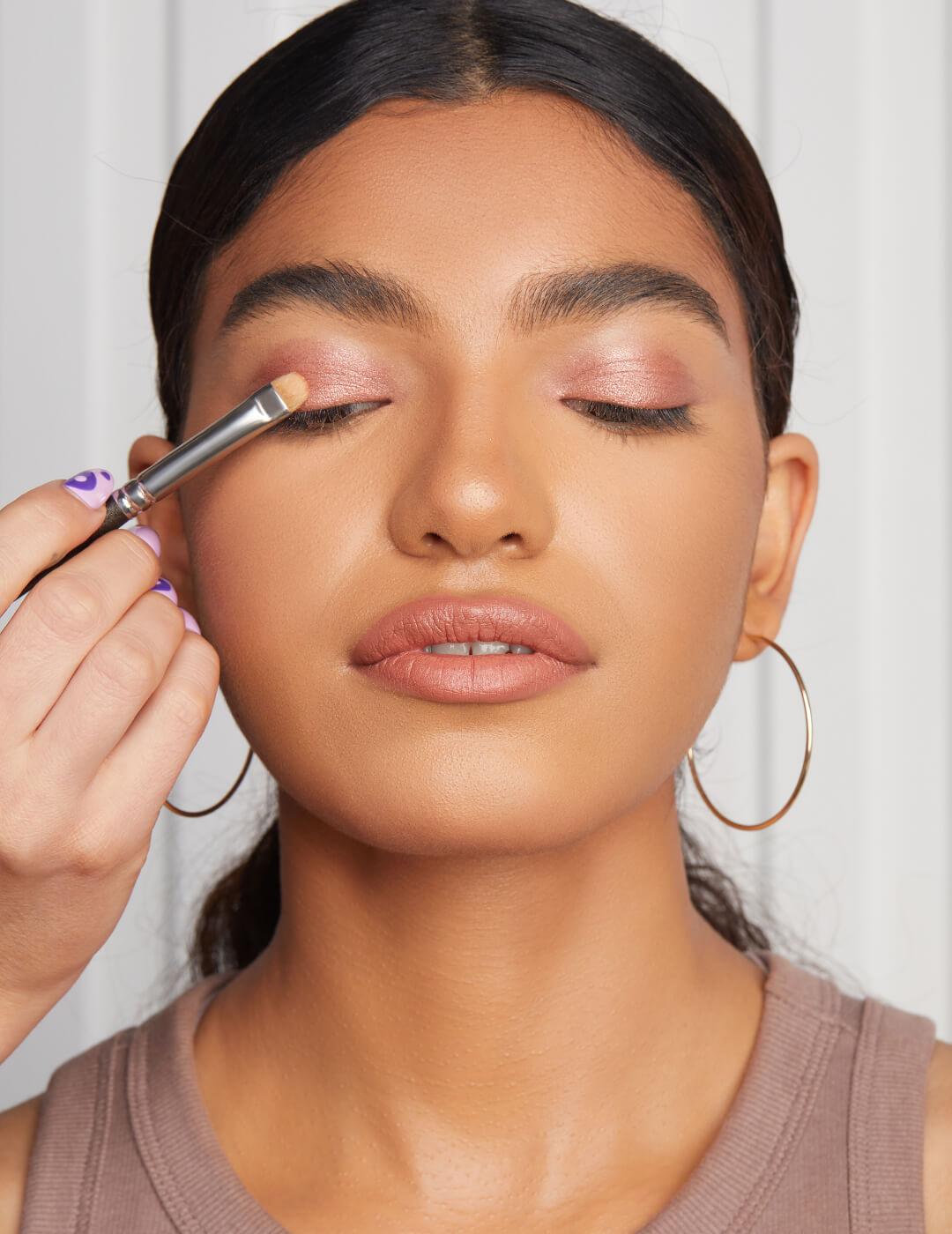 Close-up image of a makeup artist's hand using a makeup brush and applying shimmery rose gold eyeshadow on model's eyelid