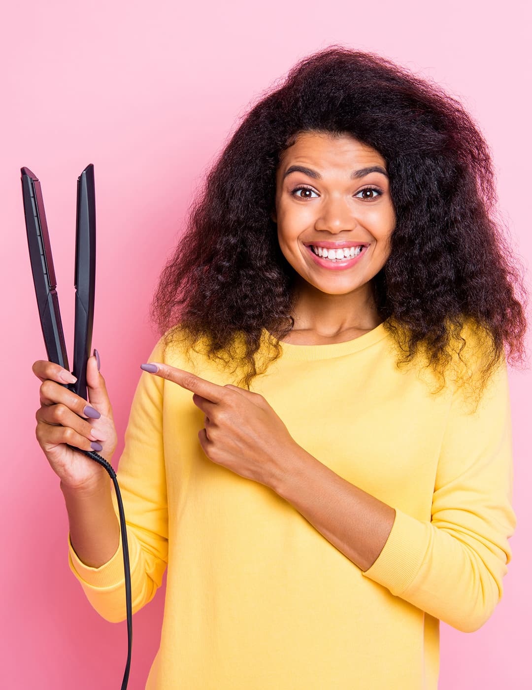 An image of a curly-haired woman showing a cheerful smile while holding a flat iron on a pink background