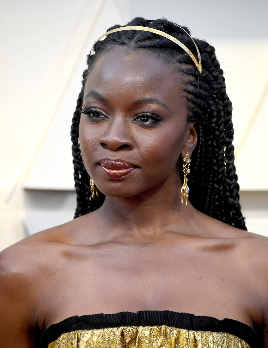 Danai Gurira looking glam in a black and gold dress, gold dangling earrings, and braided hairstyle