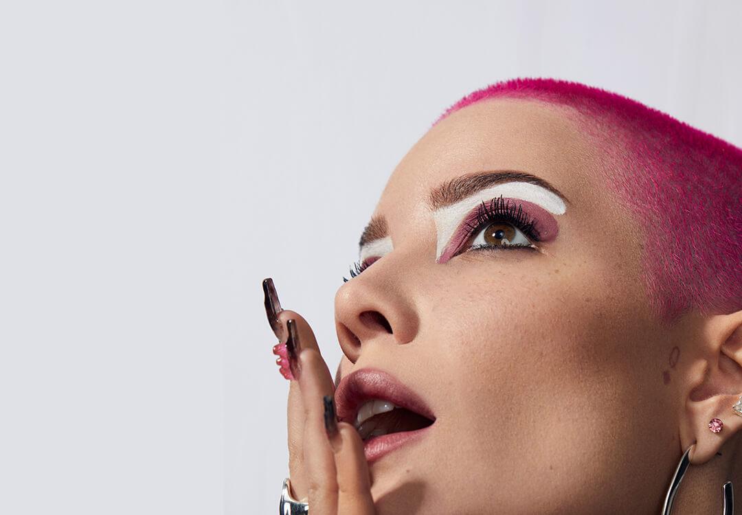 Close-up profile image of Halsey rocking bright pink hair and bold pink and white eye makeup look and posing with her hand on her mouth