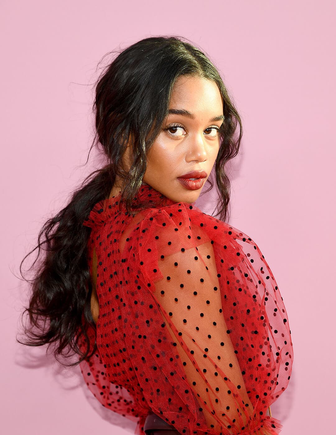 A photo of Laura Harrier sporting her black long hair in a modern romance ponytail wearing a red dress with a puffed see through sleeve in polkadots
