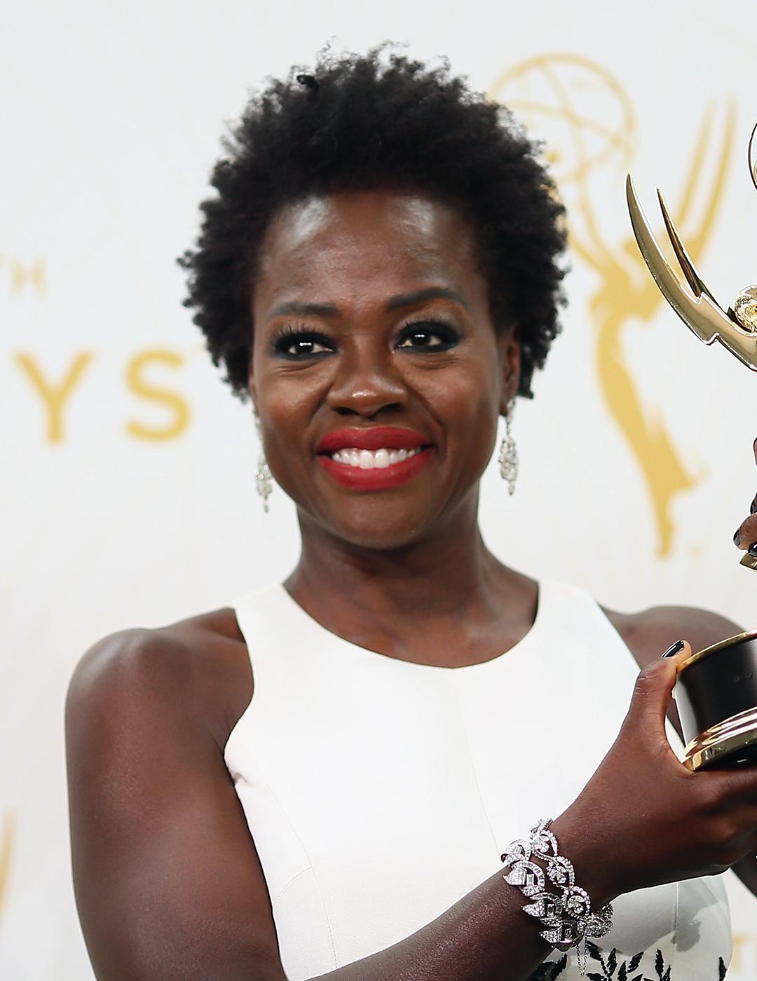 A photo of Viola Davis wearing a sleeveless white dress holding a golden trophy sporting her black short afro hair 