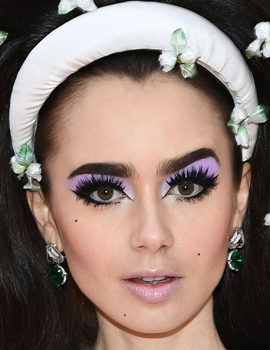Lily Collins looking retro in a purple eyeshadow makeup look, glossy and frosty nude lips, and white headband with flowers