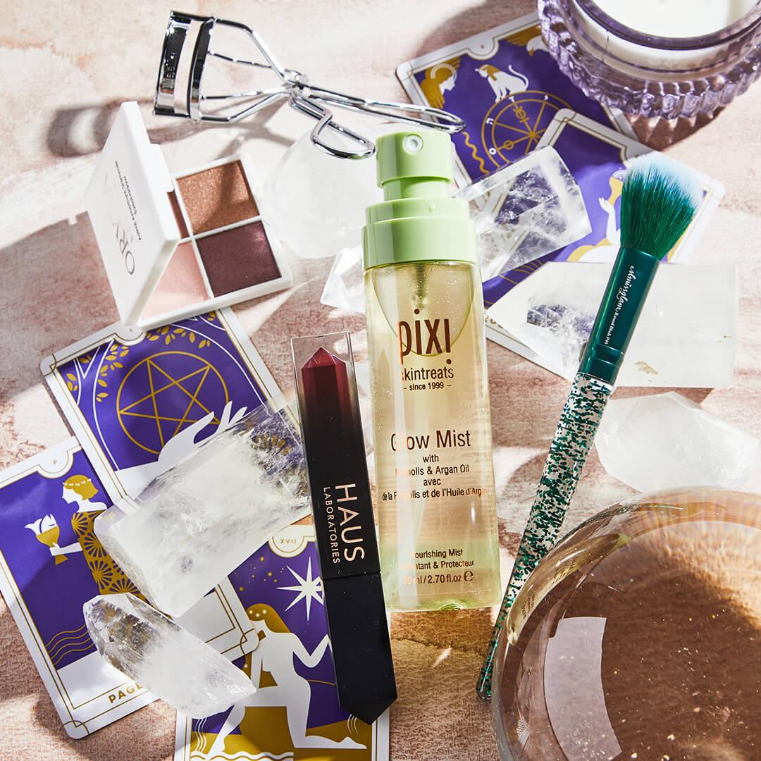 Flat lay image of skincare, makeup products and tools, crystals, and tarot cards on rose gold cloth