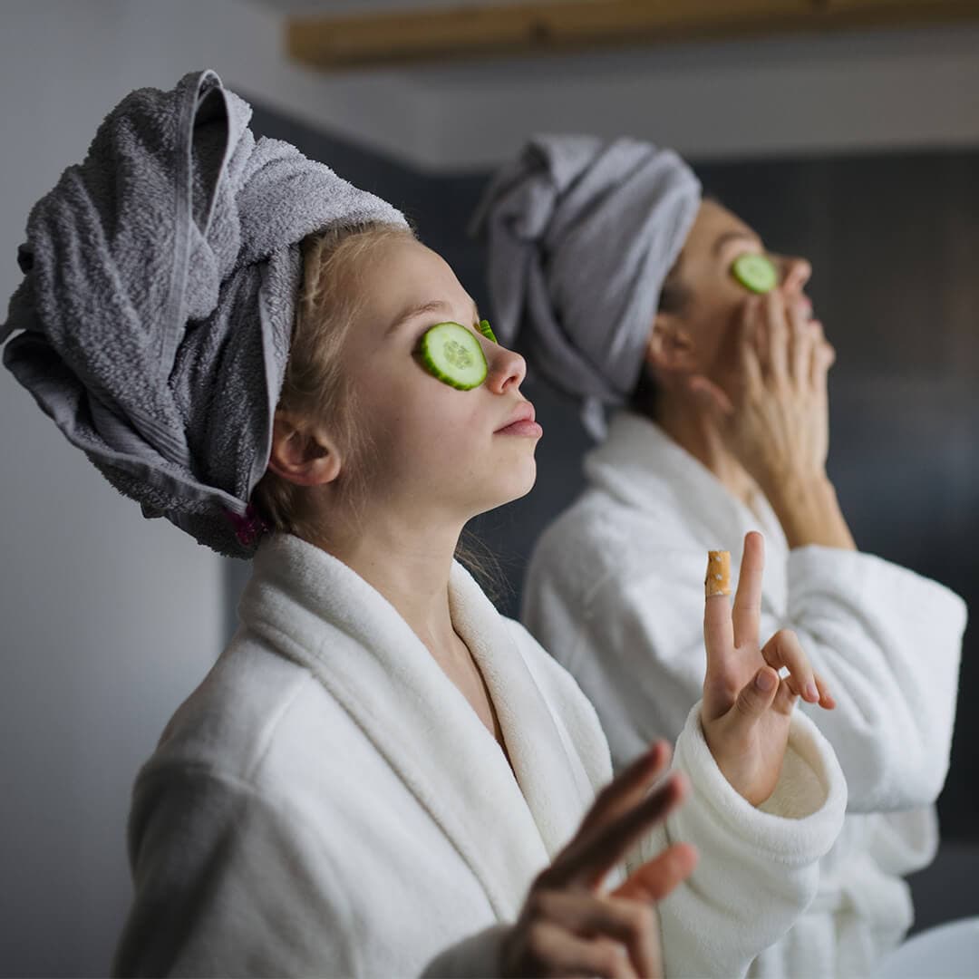 Mother and small daughter indoors in bathroom at home, putting cucumbers on eyes