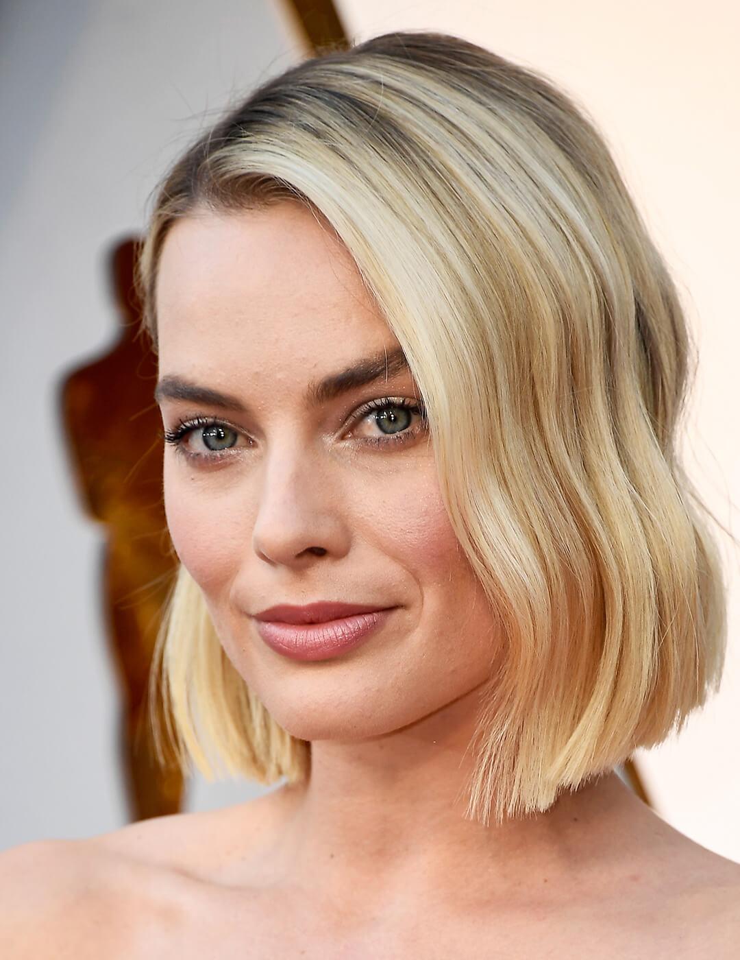 A photo of a green-eyed Margot Robbie smiling in the camera with her long bob hair style