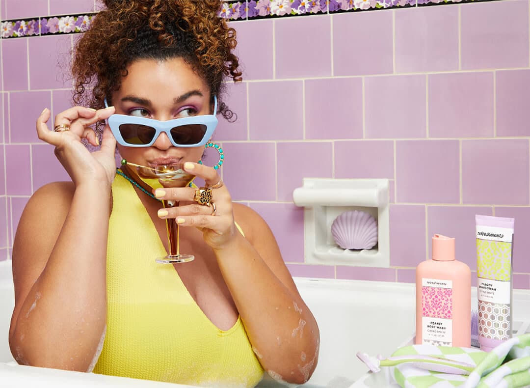Image of a curly-haired model with sunglasses sipping a cocktail in a bath tub and Refreshments personal care products beside her