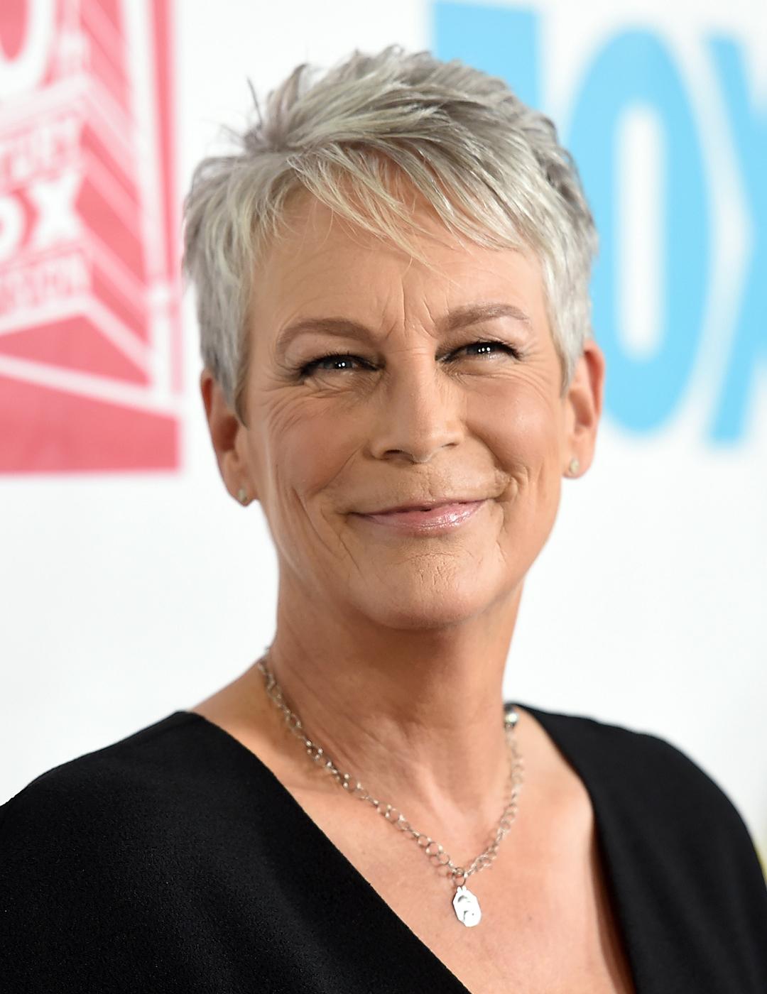 A photo of Jamie Lee Curtis wearing a plunging neckline black dress with wide sleeves and a silver necklace along with her choppy pixie haircut 