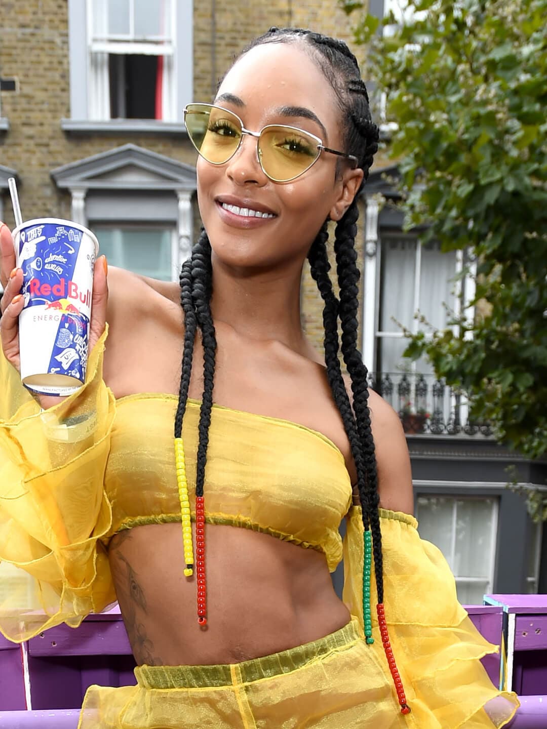 Jourdan Dunn sporting a long, braided hairstyle with colorful wraps at the end and yellow outfit while holding a can of Red Bull