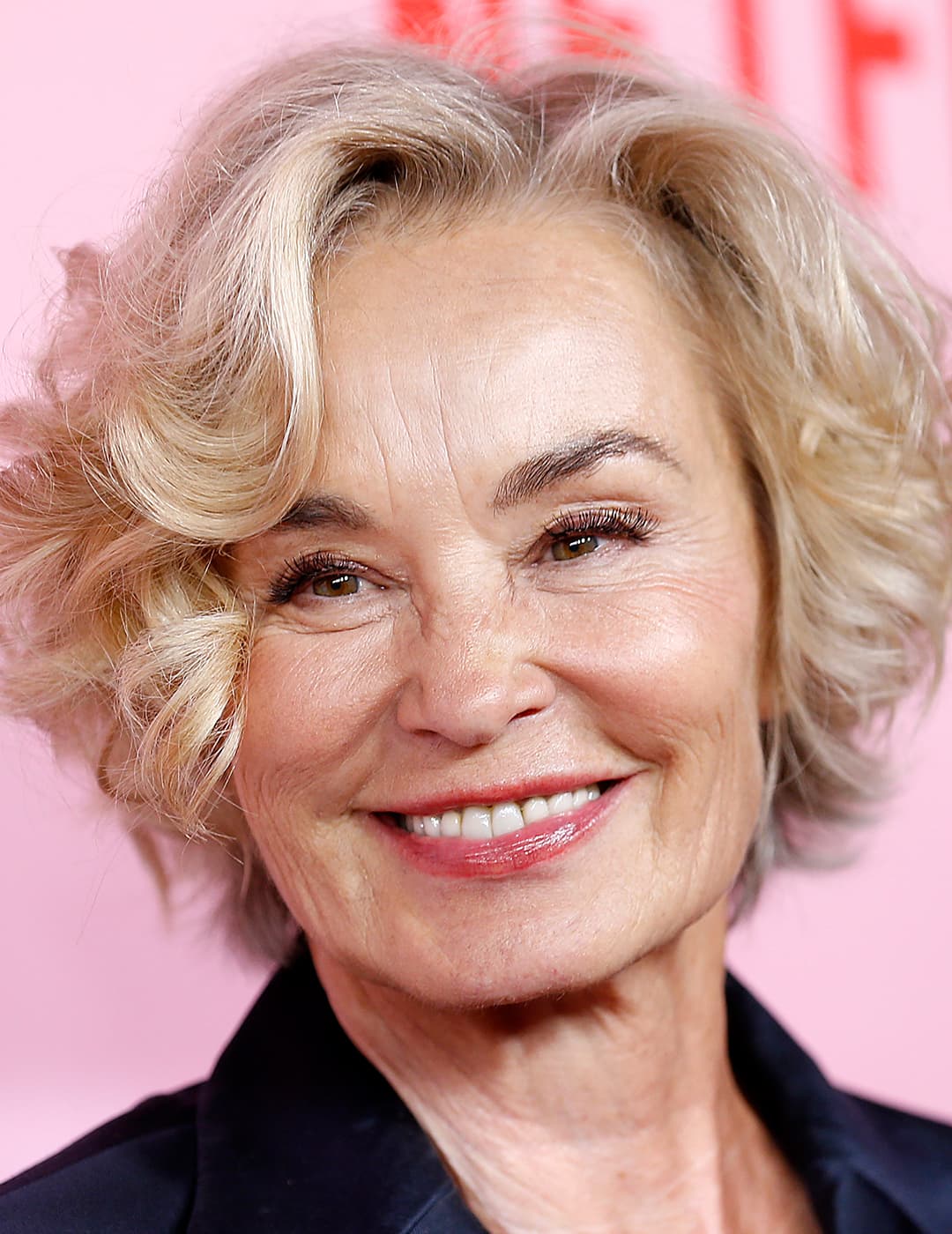 A photo of Jessica Lange in her blond angled bob cut wearing a smile and a black pant suite 