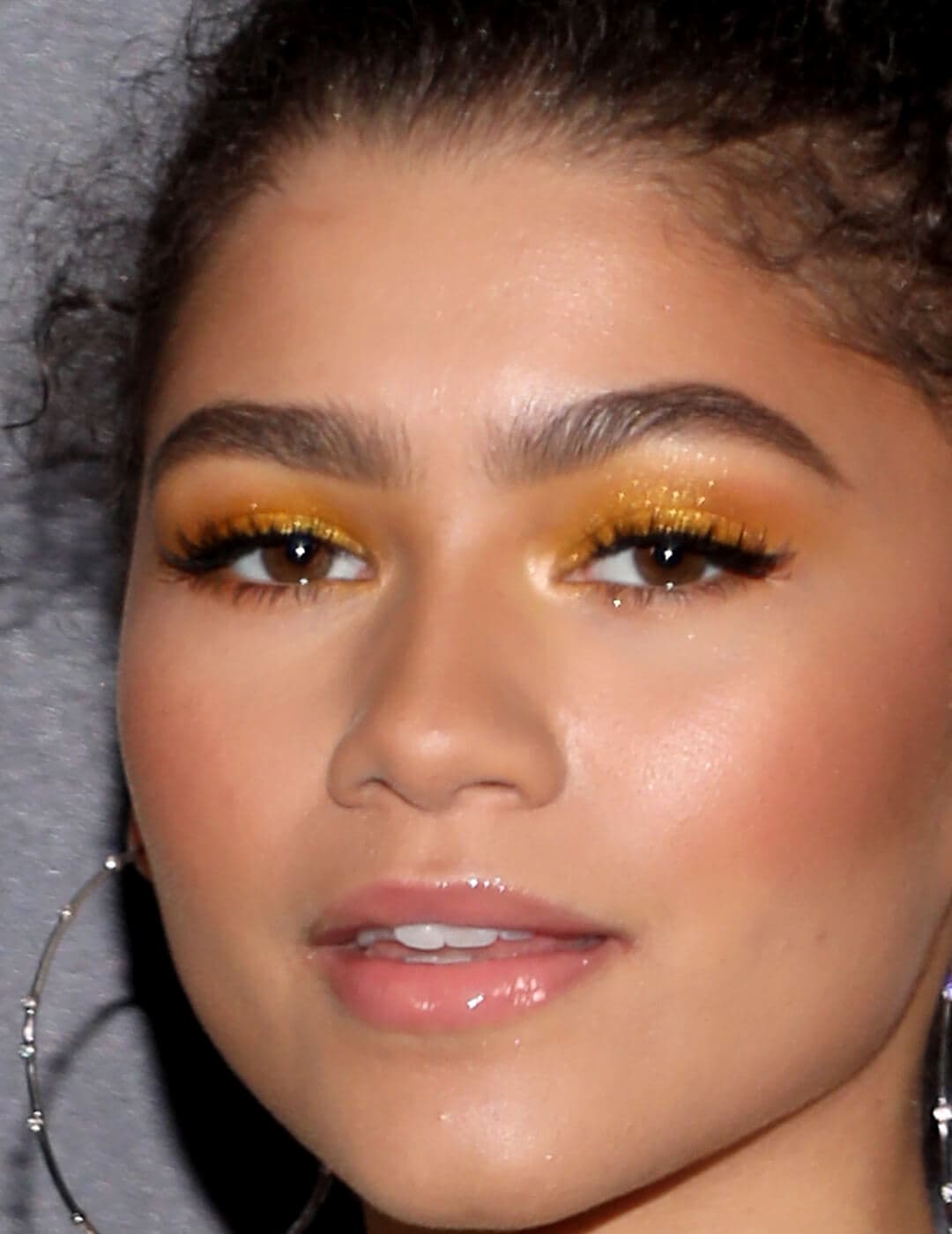Close-up image of Zendaya wearing a shimmery gold-yellow eye makeup look paired with nude lips and big hoop earrings